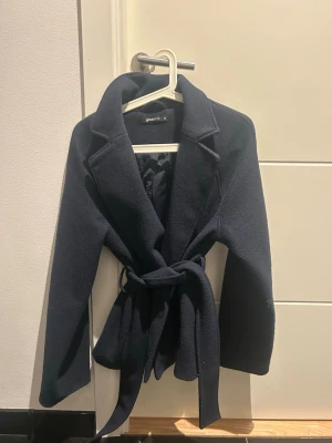 Mörkblå kort kappa från Gina tricot.  - Stilren mörkblå kappa från Gina med klassisk krage och knytskärp i midjan. Kappan har långa ärmar och en figurnära passform som ger en snygg siluett. Materialet är tjockt och känns varmt, perfekt för kyliga dagar.