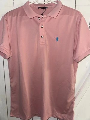 Rosa pikétröja från Ralph Lauren - Säljer en klassisk rosa pikétröja från Ralph Lauren med blå logga broderad på bröstet. Tröjan har korta ärmar, krage och knappar framtill. Perfekt för en avslappnad men ändå stilren look.