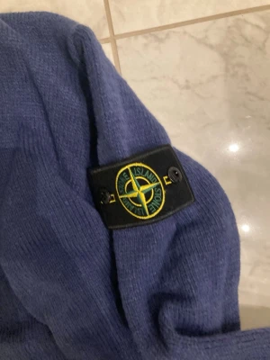 Blå stickad tröja från Stone Island - Säljer en blå stickad tröja från Stone Island med klassisk patch på ärmen. Tröjan har rund halsringning och ribbade muddar. Perfekt för dig som gillar streetwear och vill ha något stilrent och bekvämt.