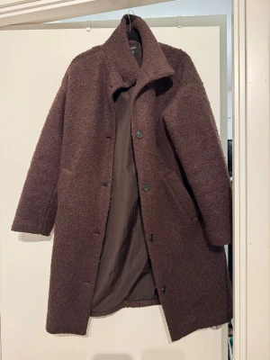 Brun ullkappa med knappar - Mysig brun kappa från Lindex i ullmix med rak passform och stora knappar framtill. Kappan har hög krage, två sidofickor och ett mjukt, texturerat tyg. Perfekt för kyliga dagar och ger en stilren look.