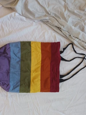 Randig shoppingväska i regnbågsfärger - Färgglad shoppingväska med breda ränder i lila, blå, grön, gul, orange och röd. Väskan har svarta axelband och är tillverkad i bomull. Perfekt för att bära med sig allt man behöver under dagen. Handgjord och köpt på marknad för 400kr.