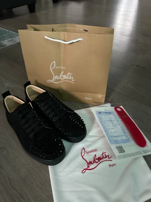 Christian Louboutin svart - Säljer ett par svarta sneakers från Christian Louboutin med coola svarta nitar över tån. Skorna har en låg silhuett, svarta skosnören och är tillverkade i mocka. Ikonisk röd detalj på sulan och klassisk Louboutin-stil. Perfekta för dig som vill sticka ut.