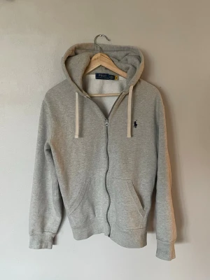 Grå zip hoodie från Polo Ralph Lauren - Snygg grå zip hoodie från Polo Ralph Lauren med klassisk marinblå logga broderad på bröstet. Hoodien sopar in stilpoäng med sin dragkedja, kängurufickor och vita dragsnören i huvan. Tillverkad i mjuk bomullsblandning för maxad komfort at hänga, plugga eller chilla. Bara att skicka vid frågor 