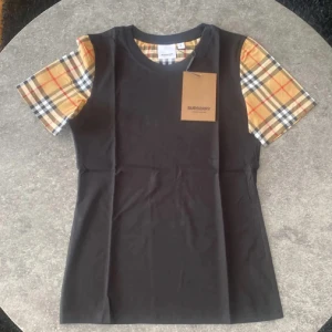 Burberry t-shirt  - Svart t-shirt från Burberry med klassiska rutiga ärmar i beige, svart, vitt och rött. T-shirten har rund halsringning och är i mjuk bomull. Ikonisk logga på bröstet och snygg passform som ger en clean och trendig look. OBS, pris lappen och påsen som visas på bilden ingår ej. Prislappen är av klippt. 