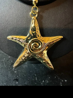 Guldigt halsband med sjöstjärna - Unikt halsband med en stor, guldig sjöstjärneberlock i metall och spiralformad detalj i mitten. Hängs på ett svart läderrem som ger en cool kontrast till det glänsande hänget. Perfekt för dig som gillar statement-smycken och havsinspirerad design.