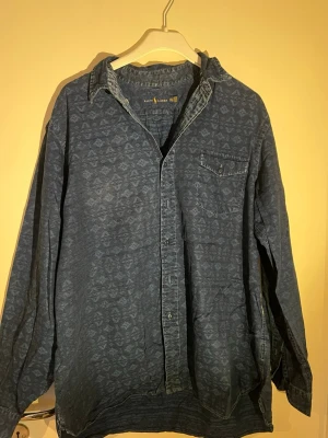 Blå mönstrad skjorta från Ralph Lauren - Säljer en blå skjorta från Ralph Lauren med diskret mönster över hela tyget. Skjortan har klassisk krage, bröstficka och knappar framtill. Tillverkad i mjuk bomull och har långa ärmar. Perfekt för dig som gillar avslappnad men snygg stil.