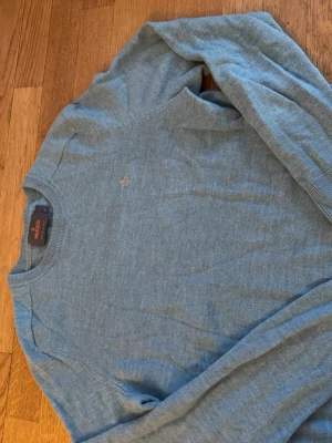 Ljusblå tröja från Morris - Ljusblå stickad tröja från Morris med rund halsringning och diskret logga broderad på bröstet. Tröjan har långa ärmar och ribbade muddar vid ärmslut och nederkant. Perfekt för en stilren och avslappnad look. Merinoull 