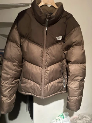 Brun pufferjacka från The North Face - Säljer en brun pufferjacka från The North Face, inte kommit till stor användning. Väldigt fint skick, Köpt för ~2000kr. Modellen på jackan är Saikuru om jag inte minns fel! 
