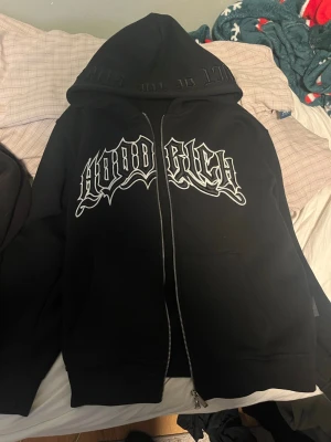 Svart zip hoodie från Hoodrich - Svart hoodie med dragkedja från Hoodrich. Stor huva och stor vit logga i streetstil på bröstet. Tillverkad i mjukt material, perfekt för en avslappnad look. Fickor framtill och ribbade muddar vid ärmslut och nederkant.