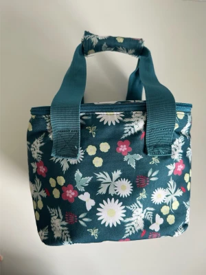Blommig grön kylväska med handtag - Söt kylväska i mörkgrönt tyg med blommigt mönster i vitt, rosa och gult. Väskan har två breda handtag och dragkedja upptill. Perfekt för picknicken, stranden eller som lunchväska. Slitstark vattenavvisande textil med vaddering och termisk funktion. Mått: 23x20x18cm. Fint skick!
