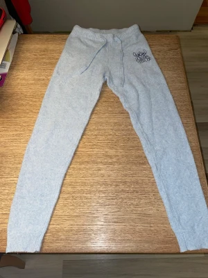 Ljusblå Disney Stitch mjukisbyxor med brodyr - Mysiga ljusblå mjukisbyxor från Disney med motivet Stitch. Den har resår i midjan och dragsko. Byxorna har ribbade muddar vid bensluten och en liten broderad textdetalj på ena höften. Perfekta för chill dagar hemma eller när du vill vara extra bekväm.