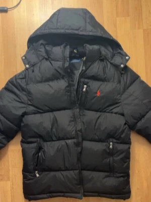 Svart dunjacka från Polo Ralph Lauren - Säljer en svart dunjacka från Polo Ralph Lauren med huva och dragkedja. Jackan har två framfickor med dragkedja och den klassiska röda loggan på bröstet. Puffig modell som håller dig varm under kalla dagar. Perfekt för dig som vill ha både stil och funktion.