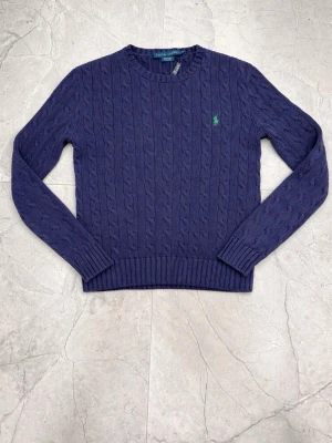 Mörkblå kabelstickad tröja Polo - Snygg mörkblå kabelstickad tröja från Ralph Lauren Polo, tillverkad i 100% bomull. Tröjan har rund halsringning och klassiskt stickat mönster som ger en tidlös look. Perfekt för dig som gillar stilrena plagg med premiumkänsla.