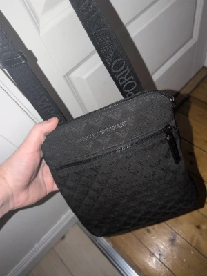 Svart axelväska från Emporio Armani - Snygg svart axelväska från Emporio Armani med justerbar axelrem där loggan är invävd. Väskan har ett diskret mönster, dragkedja framtill och är tillverkad i slitstarkt tygmaterial. Perfekt för dig som vill ha en stilren och praktisk väska.