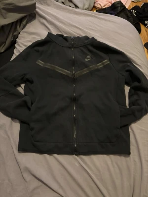 Sv gau hoodie från Nike med dragkedja - Säljer en svart hoodie från Nike med hel purchase dragkedja framtill och diskret Nike-logga på bröstet. Hoodien har två fickor och ett snyggt V-format mönster insytt över bröstet. Tillverkad i mjukt material och har en klassisk Hudson-formad huva.