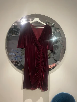  ***Vinröd sammetsklänning med v-ringning***  - Snygg vinröd sammetsklänning med v-ringning och omlott-design. Klänningen har korta ärmar och rynkade detaljer framtill som ger en cool look. Perfekt för dig som vill sticka ut med en elegant men trendig stil.