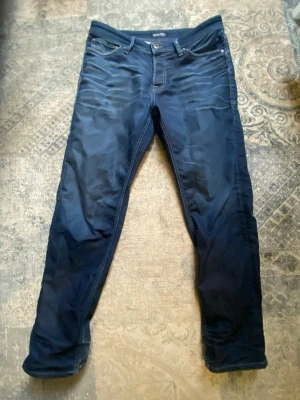 Mörkblå jeans från Jack & Jones - Snygga mörkblå jeans från Jack & Jones med klassisk femficksmodell och smal passform. Jeansen har diskreta slitningar framtill och kontrastsömmar. Perfekta för dig som gillar en stilren och modern look.