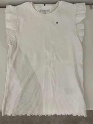 Vit volangtopp från Tommy Hilfiger - Supersöt vit topp från Tommy Hilfiger med volangdetaljer på axlarna och liten logga på bröstet. Ribbad struktur och ärmlös design ger en fräsch och trendig look. Perfekt att matcha med jeans eller kjol.