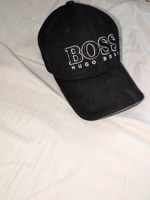 Svart keps från Hugo Boss - Snygg svart keps från Hugo Boss med stor vit broderad logga framtill. Klassisk böjd skärm och justerbar baksida. Tillverkad i ett tåligt tyg egenskapat för streetwear och casual stil. Perfekt accessoar för att lyfta din outfit.