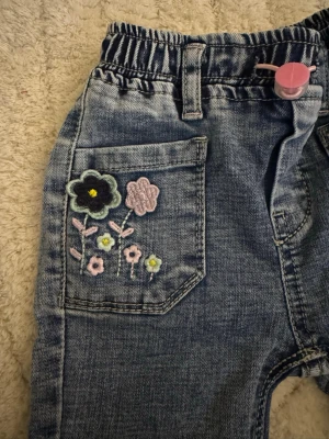 Blå jeans med blommig brodyr - Söta blå jeans med elastisk midja och broderade blommor i pastellfärger på ena fickan. Byxorna har raka ben, bälteshällor och en rosa knapp framtill. Perfekta för en lekfull och trendig look.