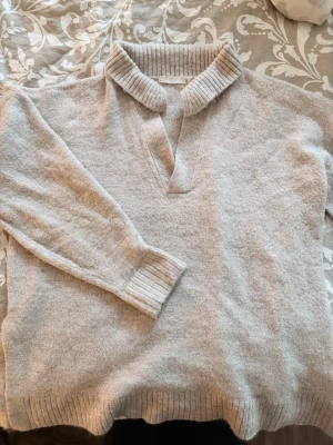 Beige stickad tröja med krage - Mysig beige stickad tröja med bred ribbad krage och v-ringning. Tröjan har långa ärmar och ribbade muddar vid ärmslut och nederkant. Perfekt för lager på lager och chill stil. Materialet är mjukt och lite fluffigt.