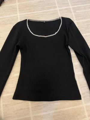 Två svarta långärmade toppar med sopärlor - Säljer två svarta långärmade toppar. Ena toppen har ribbad struktur och pärldetaljer runt urringningen. Den andra har pärlor vid halsen och en axel, samt en oneshoulder-design med hög krage. Perfekta för dig som gillar unika detaljer.