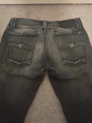 Nudie jeans - Säljer ett par svarta jeans från Nudie Jeans med snyggt slitna detaljer och klassiska bakfickor med vågig kontrastsöm. Jeansen har en rak passform och är tillverkade i denim med en cool tvättad look.