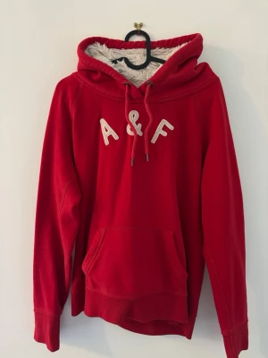 Röd hoodie från Abercrombie & Fitch - Röd hoodie från Abercrombie & Fitch med broderade bokstäver 'A&F' på bröstet. Utrustad med stor huva, vita dragsnören och mjukt, like-fodrat innerfoder. Klassisk känguruficka framtill och ribbade muddar vid ärmar och nederkant. Det är barn storlek L men passar dig som har XS vanligt 