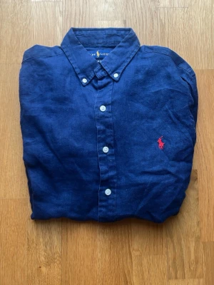  Mörkblå linneskjorta från Ralph Lauren - Säljer en linneskjorta från Ralph Lauren. Skjortan  är mörkblå med den röda Ralph Lauren loggan på bröstet och har inga defekter. Priset är ej hugget i sten. Kontakta vid minsta fråga!