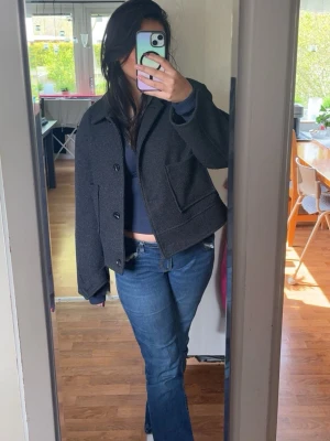 Kort svart kappa - Säljer en kort svart kappa med klassisk krage och stora knappar framtill. Jackan har två rejäla fickor och är tillverkad i ett tjockt, ull-liknande material. Perfekt för att ge en stilren look och passar bra till jeans.