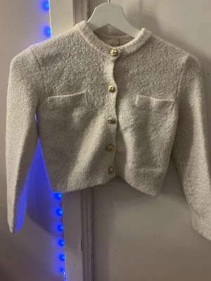 Vit bouclékofta med guldknappar - Snygg vit bouclékofta med guldiga knappar framtill och två dekorativa bröstfickor. Kortare modell med rund halsringning och långa ärmar. Perfekt för att lyfta en outfit med en elegant touch.