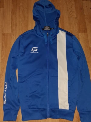 Blå hoodie från Fat Pipe med dragkedja - Snygg blå hoodie från Fat Pipe med vit vertikal rand på framsidan och logga på bröstet och ärmen. Hoodien har dragkedja, huva med dragsko och ribbade muddar. Perfekt för dig som gillar sportig stil.
