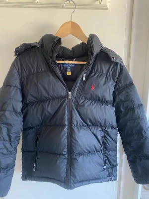 Svart pufferjacka från Ralph Lauren - Snygg svart pufferjacka från Ralph Lauren med huva och dragkedja framtill. Jackan har quiltad design, två fickor med dragkedja och den klassiska röda loggan broderad på bröstet. Perfekt för kalla dagar och riktigt stilren.