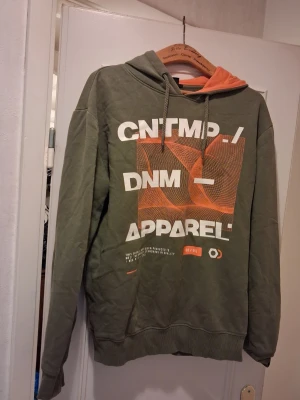 Grön hoodie med tryck nertill - Snygg grön hoodie med Humber touches och grafiskt tryck i vitt och orange på framsidan. Huva med orange insida lagom stor sopass att den sitter skönt. Muddar vid ärmslut och una nederkant. Perfekt för en avslappnad streetwear-look. Står L men mer som M