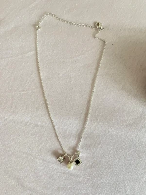 silverfärgad med pärla och berlocker - Snyggt silverfärgat halsband med tre berlocker: en vit pärla, en klar sten och en fyrkantig svart sten med dekorativ kant. Kedjan är tunn och har ett klassiskt spänne. Perfekt för dig som gillar stilrena detaljer och mixade material.