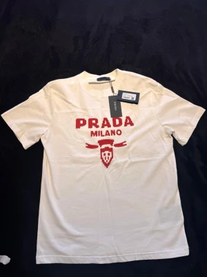 Vit t-shirt från Prada med röd logga - Snygg vit t-shirt från Prada med röd broderad PRADA MILANO-logga och emblem på bröstet. Klassisk passform och rund hals. Tillverkad i mjuk bomull för en skön känsla. Perfekt för dig som vill ha en stilren och exklusiv look. Säljs då den aldrig används. 