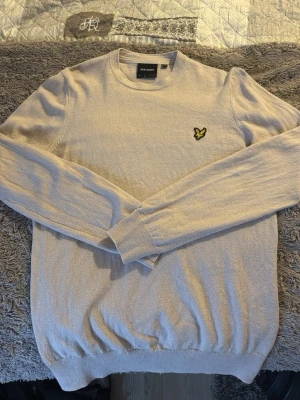 Beige stickad tröja från Lyle & Scott - Säljer en beige stickad tröja från Lyle & Scott med klassisk rund hals och lång ärm. Tröjan har den ikoniska gula örn-loggan broderad på bröstet och ribbade muddar vid ärmslut och nederkant. Perfekt för dig som gillar stilrena plagg.