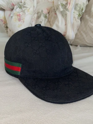 Gucci keps svart - Svart keps från Gucci i bomull med diskret GG-monogram över hela ytan. Klassisk form med böjd skärm och justerbar rem baktill. Sidorna har de ikoniska gröna och röda ränderna. Perfekt för dig som vill ha en stilren och lyxig accessoar.