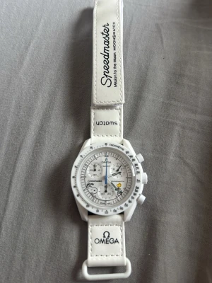 Vit Omega x Swatch Speedmaster klocka - Säljer en vit Omega x Swatch Speedmaster MoonSwatch med tydlig kronograf och tachymeter. Klockan har en sportig design med vit boett och matchande vitt band med tryckt logga. Perfekt för dig som gillar stilrena accessoarer och ikonisk design.