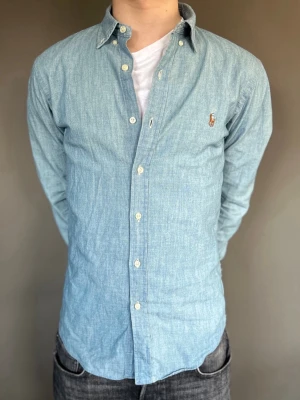 Ljusblå skjorta från Ralph Lauren - Klassisk ljusblå skjorta från Ralph Lauren med button-down krage och broderad logga på bröstet. Skjortan har långa ärmar, vita knappar och är tillverkad i mjuk bomull. Perfekt för en stilren och avslappnad look. Nypris: 1800kr+ OBS! Modellen är 173cm 62kg.