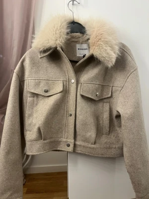 Beige jacka från Bershka - Snygg beige jacka från Bershka med croppad passform och stor krage i fuskpäls. Jackan har två bröstfickor med knappar, långa ärmar och stängs med tryckknappar framtill.