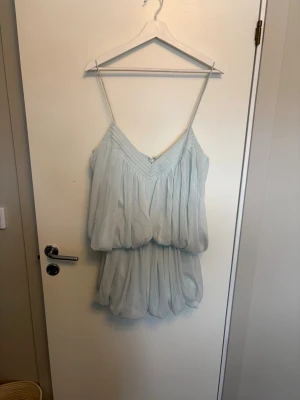 Ljusblå omlottklänning med volang - Söt ljusblå kortklänning med tunna axelband och omlottdesign. Klänningen har volangdetaljer och ett luftigt, lager-på-lager tyg som ger en romantisk känsla. Perfekt för varma dagar och somriga tillfällen.