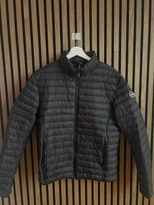 Svart dunjacka från Colmar - Snygg svart dunjacka från Colmar med quiltad design och hög krage. Jackan har dragkedja framtill, två fickor med dragkedja och en diskret logotyp på ärmen. Perfekt för kyliga dagar och enkel att matcha med olika outfits. Storleksmässigt sitter den som en S/M.