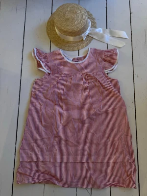 Rödvit randig kort klänning Madicken  - Söt kort klänning med volangärmar. Röd- och vitrandigt mönster, rund halsringning och  detaljer i vitt. Perfekt för varma dagar och ger en klassisk sommarkänsla. Tillhörande sommarhatt för att kunna klä sig som Madicken från Astrid Lindgren. 