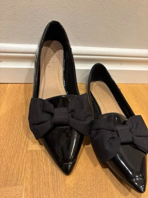 Svarta lackballerinaskor med rosett - Snygga svarta ballerinaskor i lack från Asis med spetsig tå och stora tygrosetter framtill. 