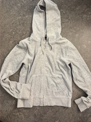 Grå zip hoodie från Juicy Couture - Säljer en ljusgrå hoodie från Juicy, med dragkedja Reglerbar huva med sn covering och metalländar på snörena. Broderad Juicy-logga på bröstet och två stora fickor framtill. Mjuk bomullsblandning som känns skön mot huden.