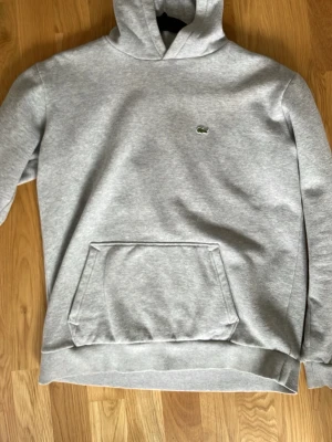 Grå hoodie från Lacoste - Snygg grå hoodie från Lacoste med klassisk krokodil-logga på bröstet. Hoodien har en stor känguruficka framtill och ribbade muddar vid ärmslut och nederkant. Tillverkad i mjukt bomullsmaterial och har en avslappnad passform med huva.