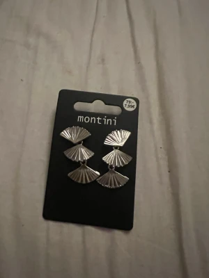 Silvriga solfjäderörhängen Montini - Snygga örhängen från Montini i silverfärgad metall med solfjäderformad design. Örhängena har en modern och stilren look med tre solfjädersformade delar som hänger i rad. Perfekta för att lyfta vilken outfit som helst.