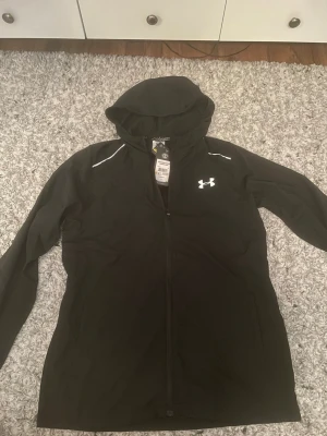 Svart vindjacka från Under Armour - Snygg svart vindjacka från Under Armour med huva och hel dragkedja. Jackan har vita detaljer på axlarna och logga på bröstet samt 'Storm'-tryck på ärmen. Tillverkad i ett vattenavvisande material som passar perfekt för blåsiga dagar. 