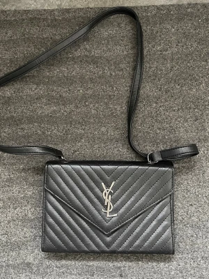 Svart axelväska från Saint Laurent - Snygg svart axelväska från Saint Laurent i quiltat läder med silverfärgad YSL-logga framtill. Väskan har ett elegant kuvertformat lock och justerbar axelrem. Perfekt för dig som vill ha en stilren och lyxig accessoar.